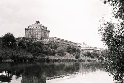 A large, industrial complex sits atop a man-made embankment, overlooking a calm river. The building features a central tower ...