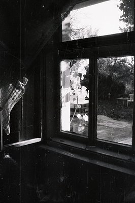 A vintage black and white photograph captures a view through a window, showing a garden scene. A loosely knit shawl hangs on ...