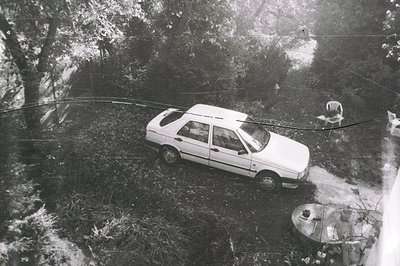 High-angle, monochrome image depicts a white sedan parked on a dirt path through a densely wooded area. A round table with di...