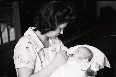 A woman in a short-sleeved, button-down gown gently cradles a sleeping infant in a hospital setting. Strong directional light...