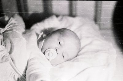 Infant lies asleep, pacifier in mouth, swaddled in a textured blanket. Appears to be a candid, intimate moment, likely a fami...