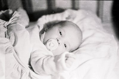 Infant rests on textured bedding, holding a pacifier. Close-up captures a direct gaze, suggesting a candid moment. Likely a f...