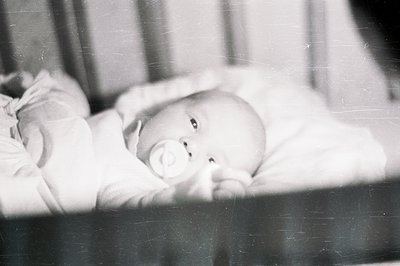 A baby lies in a crib, clutching a pacifier. The monochrome image suggests a candid, personal snapshot, likely from the mid-2...