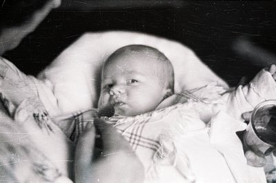 Close-up, black & white portrait of an infant cradled in arms. Baby wears a delicate, ruffled dress. Grainy texture & visible...