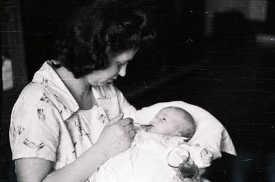 A young woman in a floral print dress tenderly feeds a baby from a bottle. The image, likely a candid moment, displays a clos...