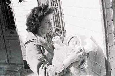 A woman, possibly a mother, holds a swaddled infant outside a building, likely a home. Architectural details suggest an early...