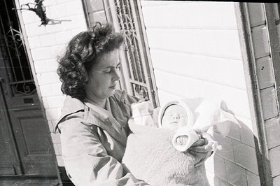 A young woman holds a swaddled baby on a porch with ornate wrought-iron details. Likely taken in the 1940s or 50s, it portray...