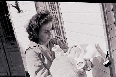 A young woman holds a bundled infant, likely a newborn, in a brick-walled exterior. Visible architectural details suggest a r...