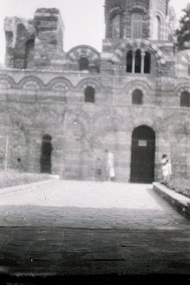 Monochrome photograph depicting the facade of a stone building, likely a church or monastery. Arched windows punctuate the st...