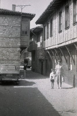 A young boy and woman walk down a narrow, cobblestone street lined with traditional timber-framed buildings. A car (license p...