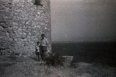 Two figures stand near a rough-hewn stone wall overlooking the sea. The wall appears to be part of an older structure, likely...