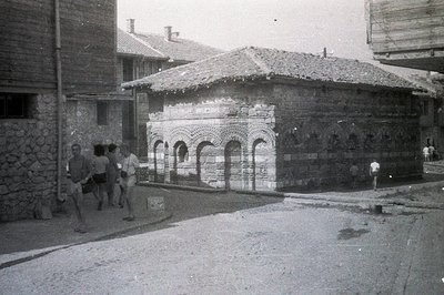 A unique, compact building with arched brickwork and a low, tiled roof stands on a dusty, cobbled street. Three men in swimwe...
