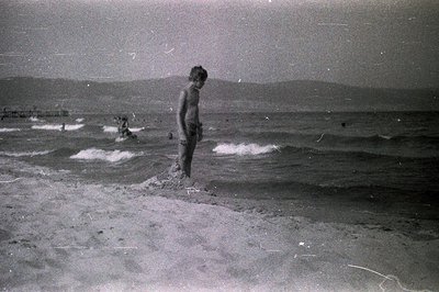 A young, barefoot boy stands on a sandy beach facing the sea, possibly contemplating the waves. Distant figures and a structu...