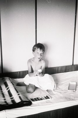 A young boy, shirtless and wearing only shorts, sits on a bed holding a small rectangular object, likely a photograph. The ro...