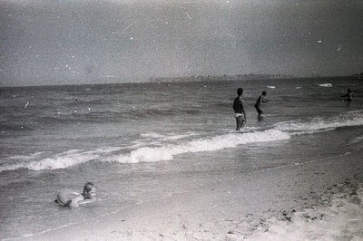 Seascape view features two boys wading in the shallows, a third person swimming, and a distant coastline. The grainy, vintage...