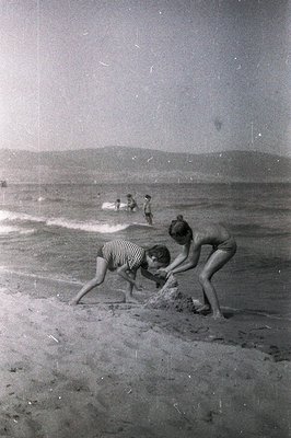 Two children building a sandcastle on a wide beach; figures in the distance. Soft focus, suggesting a candid snapshot. Likely...