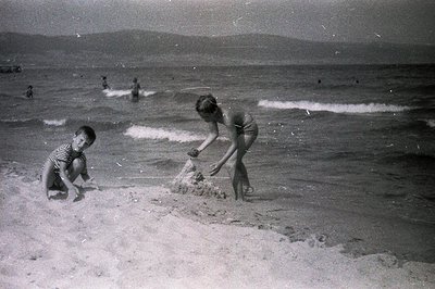 Two children build a sandcastle near the water's edge on a crowded beach, framed by distant hills. The girl wears a one-piece...