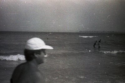 A blurry, candid monochrome captures a coastal scene. A figure in a baseball cap stands near the water’s edge, partially obsc...