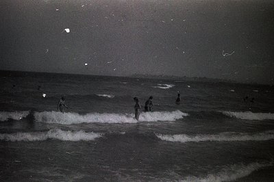 A black and white photograph depicts figures wading through breaking waves at night. The scene, likely a seaside location, sh...