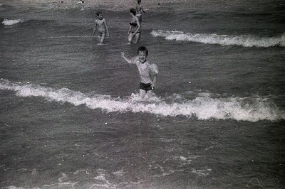 A black and white image depicts three young boys playing in the surf. The central boy, mid-stride, raises his arm in a wave. ...