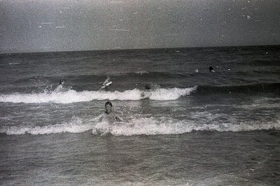 A monochrome photograph shows several figures wading through breaking waves in a coastal setting. The image captures motion a...