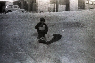 A child in winter clothing, including a hooded hat, crouches on a snow-covered slope, apparently operating a primitive sled. ...