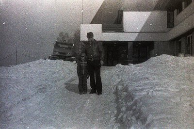 A snowy scene depicts two figures in heavy coats standing in front of a modernist building. Deep snowdrifts dominate the fore...
