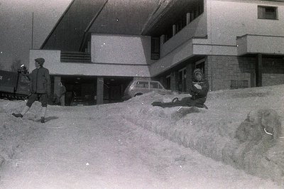 Monochromatic scene depicts a modern, concrete building with an overhanging design. A man in a hat strides across a large sno...