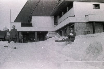 A man in a coat stands in deep snow, observing a child sliding down a large snowbank. The backdrop features a modernist build...