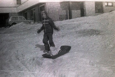 A young child, bundled in a snowsuit and hat, stands next to a wooden toboggan on a snow-covered slope. A vintage car is visi...