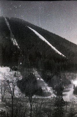 Monochrome landscape captures a snow-covered hillside with a marked ski run. Dense evergreen forest flanks the slope, leading...