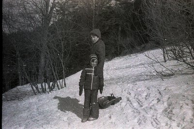 A young boy stands on a snow-covered slope, bundled in a thick winter coat, hat, and snow pants. A wooden toboggan rests at h...