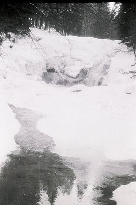 Monochrome landscape depicting a partially frozen stream flowing through a snow-covered forest. The water reflects dark tree ...