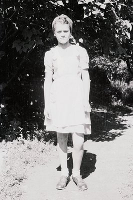 A young girl stands on a stone path, wearing a simple, knee-length dress and dark shoes with socks. Her hair is styled in sho...