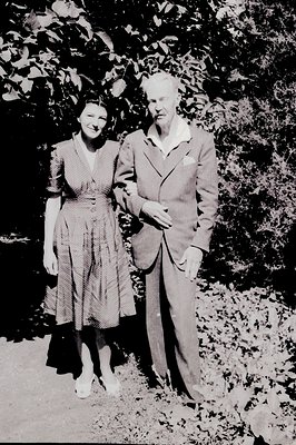 A posed portrait of a young woman and older gentleman, likely a couple, standing outdoors among foliage. Woman wears a knee-l...