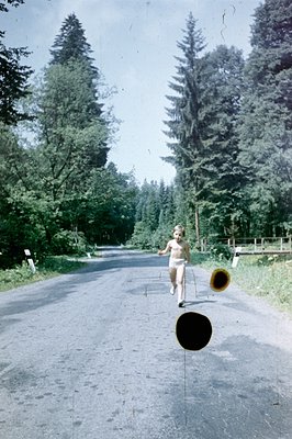 A young child runs along a paved road through a dense, coniferous forest. The scene appears rural, possibly in an alpine sett...