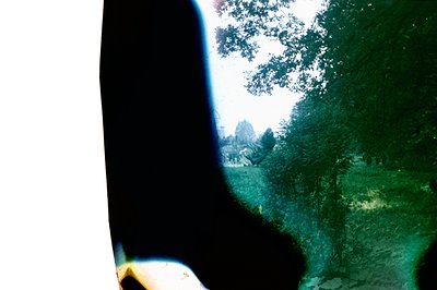 A vintage color photograph shows a view through foliage, revealing a building and a green lawn. The image exhibits a warm col...