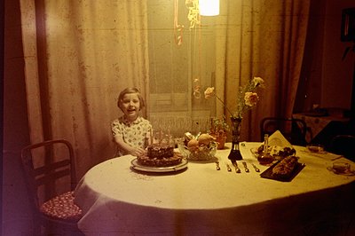 A young girl sits at a formally set table, a multi-tiered birthday cake prominently displayed. The room's décor suggests a mi...