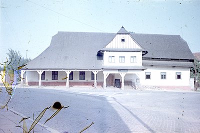 A two-story building, likely a train station or public hall, with a steeply pitched, slate roof and a covered, open porch. Th...