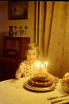 A young girl sits at a table, beaming at a layered cake with lit candles. Likely a birthday celebration, the room features fl...