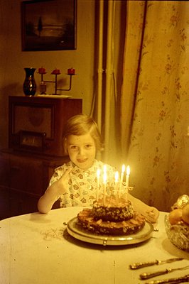 A young child, possibly a girl, smiles warmly at the camera, seated at a table with a layered cake adorned with lit candles. ...