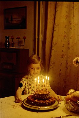 A young boy with short hair smiles while seated at a table with a cake topped with six lit candles. Floral wallpaper and patt...