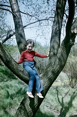 A young girl in a red turtleneck and bell-bottom jeans playfully perches in a tree. She displays a joyful expression. Likely ...