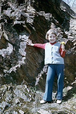 A young girl, circa 1970s, stands before a textured rock face, wearing a red turtleneck, blue bell-bottoms, and a quilted ves...