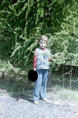 Young child stands on a gravel path, holding a black disc. Wearing a red long-sleeved shirt, blue vest, flared jeans, and whi...