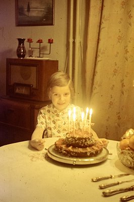 A young child sits at a table, smiling at a cake adorned with lit candles. The room features a floral wallpaper, vintage furn...