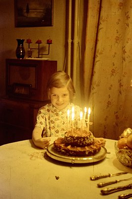 Young girl seated at a table with a layered cake, adorned with lit candles. The room features floral wallpaper, a dark wood c...