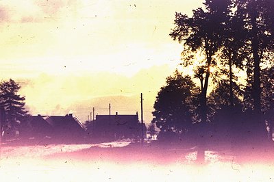 A pastel-toned image shows a rural landscape with silhouetted trees and buildings against a colorful, hazy sky, likely a suns...