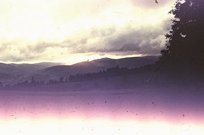 Dramatic cloudscape over a forested landscape. A body of water reflects the sunset, tinted in pink and purple hues. The scene...