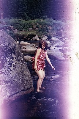 A woman in a red, short-sleeved floral dress steps across a shallow, rocky stream. Lush greenery frames the water, indicating...
